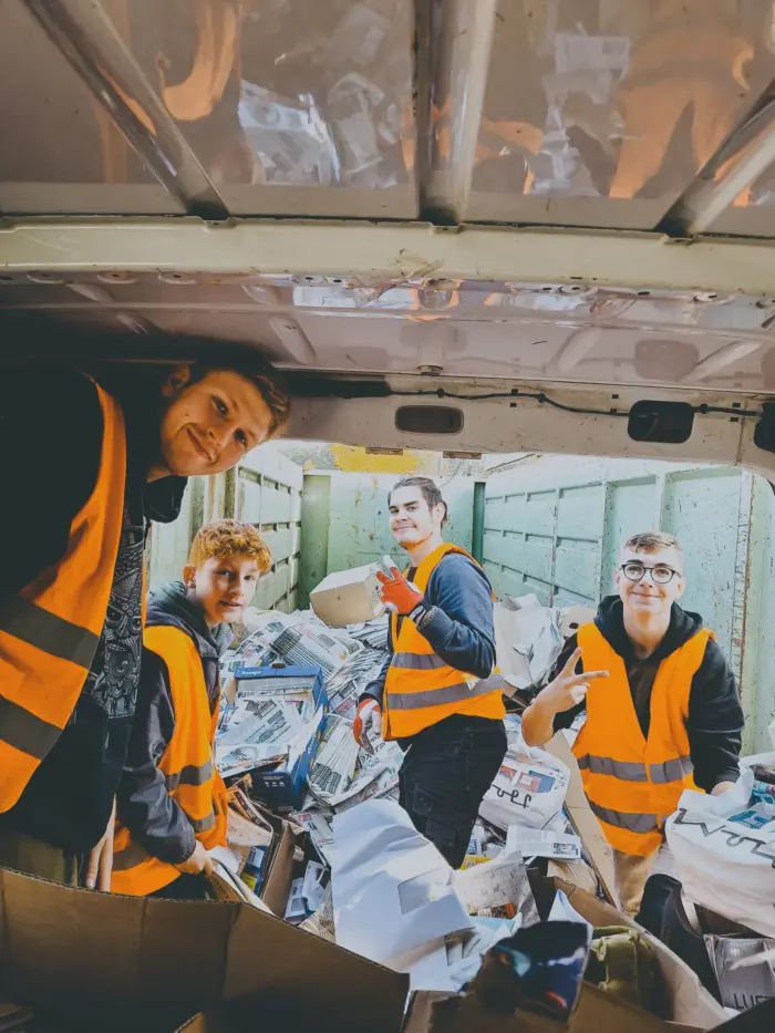 Vier Jugendliche in orangen Warnwesten sortieren lachend Zeitungen auf der Ladefläche.
