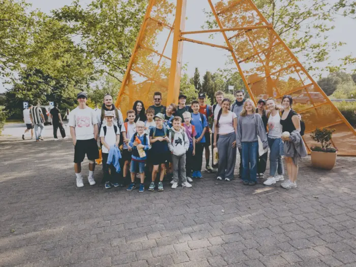 Gruppenfoto der Pfarrjugend mit Kindern und Leitern beim Ausflug in den Skylinepark, vor einer orangen zeltförmigen Stahlskulptur.