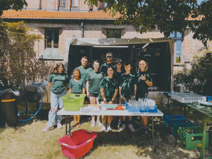 Neun Jugendliche in grünen Vereins-T-Shirts stehen bei Sonnenschein vor dem Küchenanhänger am Spültisch.