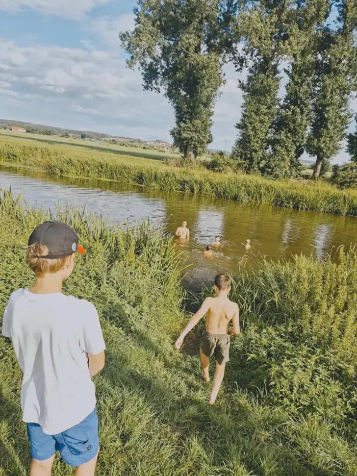 Jungen baden in einem Fluss, ein weiterer läuft durch hohes Gras zum Ufer hinunter.