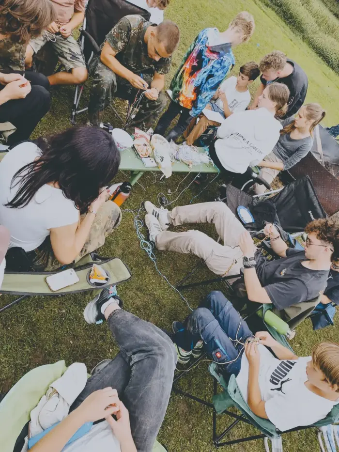 Jugendliche sitzen im Kreis auf Campingstühlen auf einer Wiese und knüpfen gemeinsam Freundschaftsbänder.