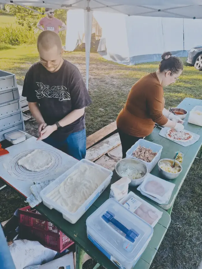 Zwei Jugendliche formen und belegen Pizzateig an einer Biertischgarnitur im Abendlicht des Zeltlagers.