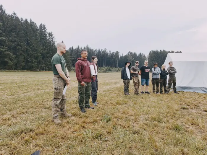 Eine Gruppe steht auf der Lagerwiese vor einem großen weißen Zelt und hört einem Betreuer zu.