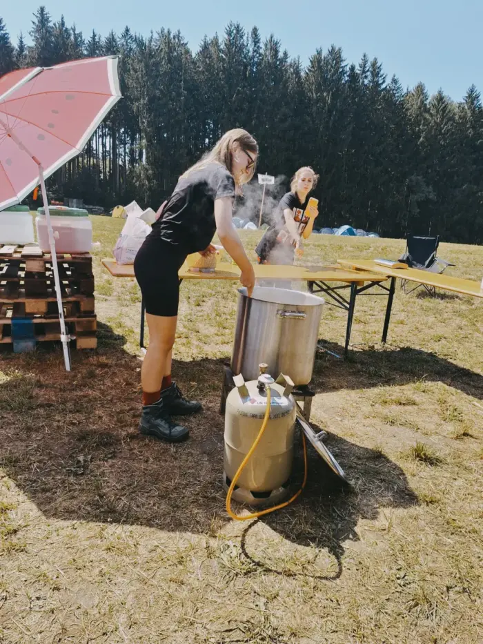 Eine Jugendliche rührt in einem großen Edelstahltopf auf einem Gaskocher, daneben ein Campingtisch unter einem Sonnenschirm.