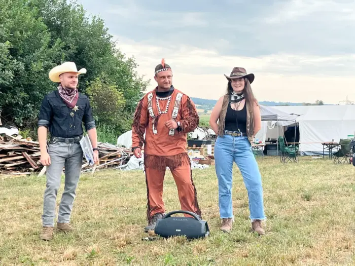 Dieselben drei in Wildwest-Verkleidung, näher fotografiert, vor Wiese und Lagerzelten.