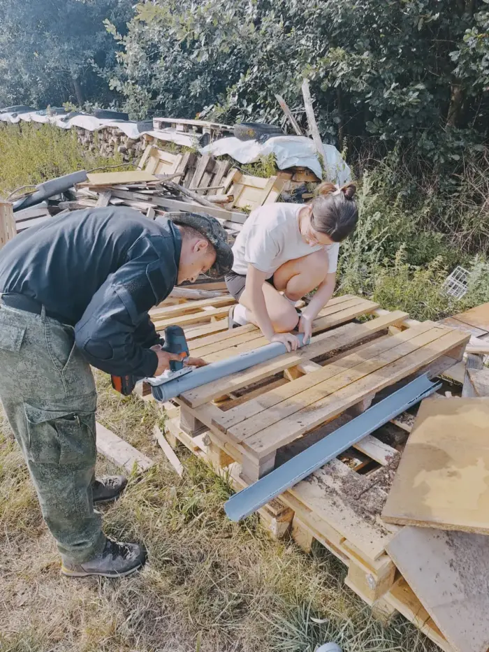 Zwei Jugendliche schrauben auf einer Wiese Holzpaletten zusammen, einer setzt den Akkuschrauber an.