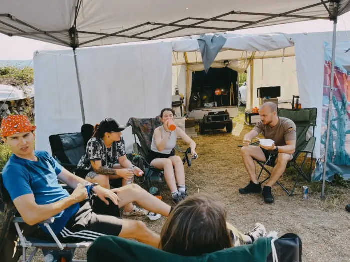 Jugendliche sitzen in Campingstühlen unter einem Pavillon neben dem Zelt und trinken etwas.