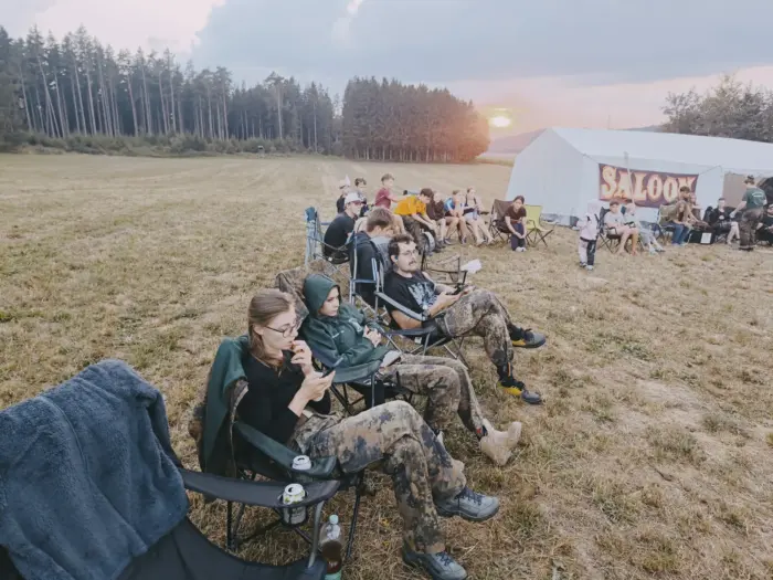 Jugendliche in Campingstühlen auf der Lagerwiese, dahinter ein großes weißes Zelt mit der Aufschrift Saloon vor untergehender Sonne.