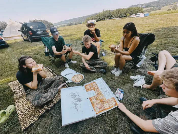 Eine Gruppe sitzt und liegt auf Decken im Gras, isst Pizza und blättert in einem großen bemalten Buch.