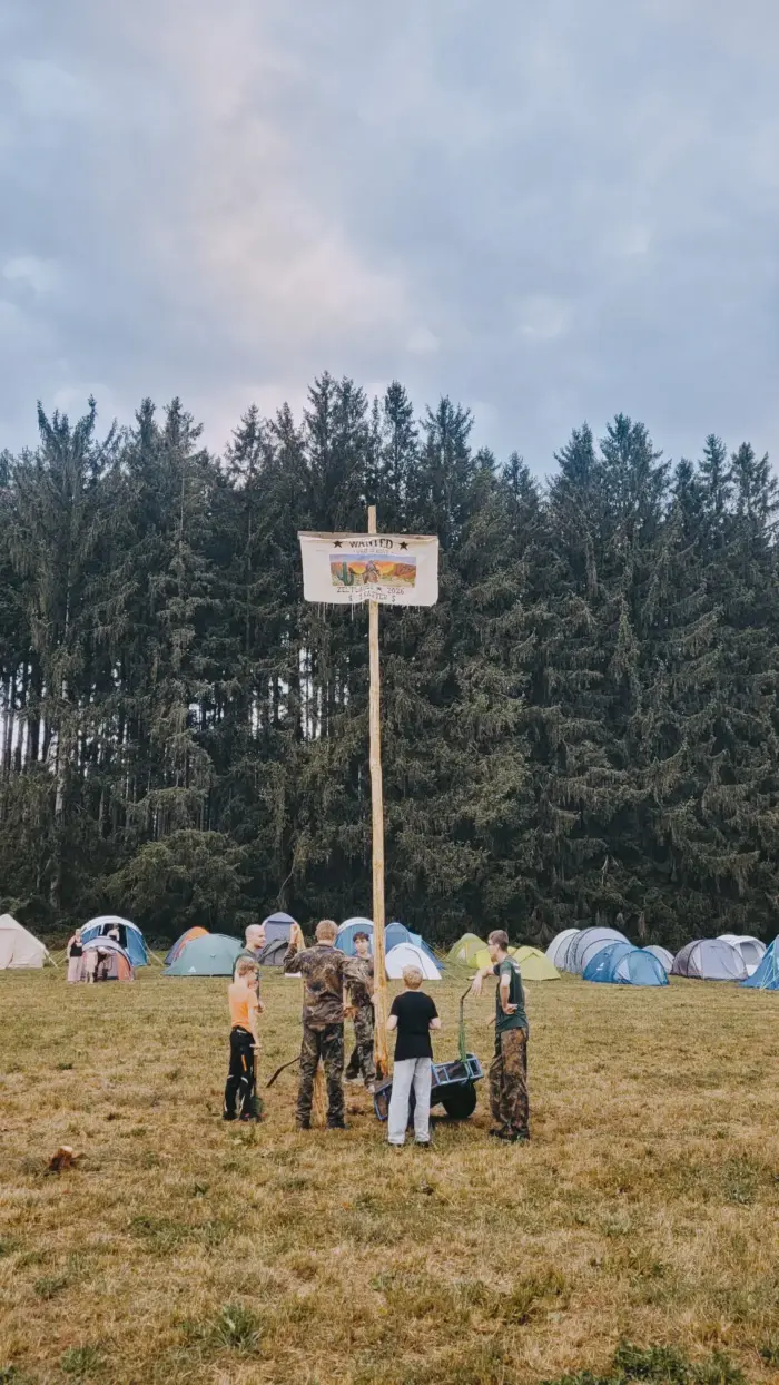Ein hoher hölzerner Wegweiser mit bemaltem Schild auf einer Wiese, dahinter das Zeltdorf und ein Nadelwald.